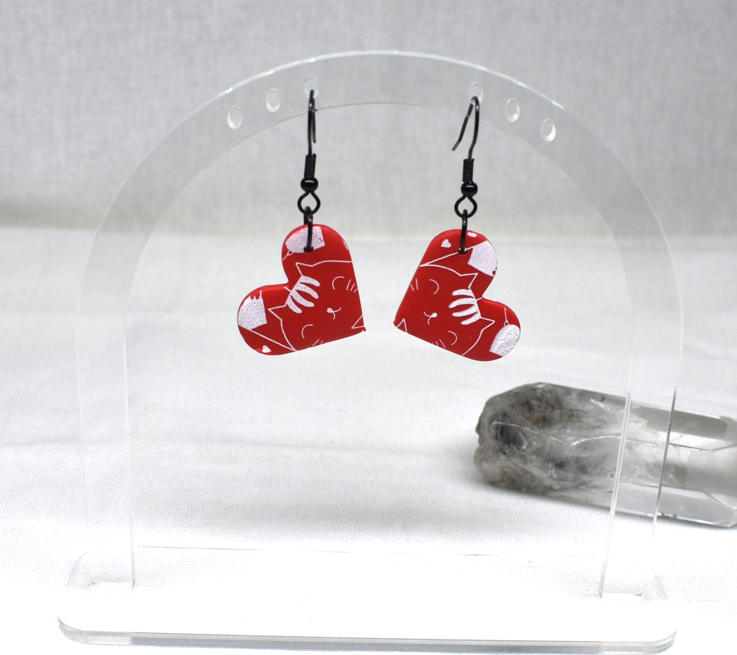 Red heart-shaped acrylic dangle earrings with white cat illustrations, displayed on a clear acrylic stand next to a clear quartz crystal.