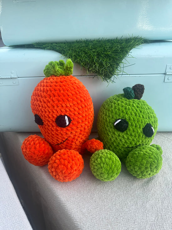 Two cute crocheted plush toys shaped like a carrot and an apple, sitting side by side on a light gray fabric surface in front of a white suitcase with artificial grass.