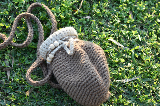 A handmade crocheted shoulder bag in taupe brown with a cream-colored beaded trim and strap, resting on a green grass background.