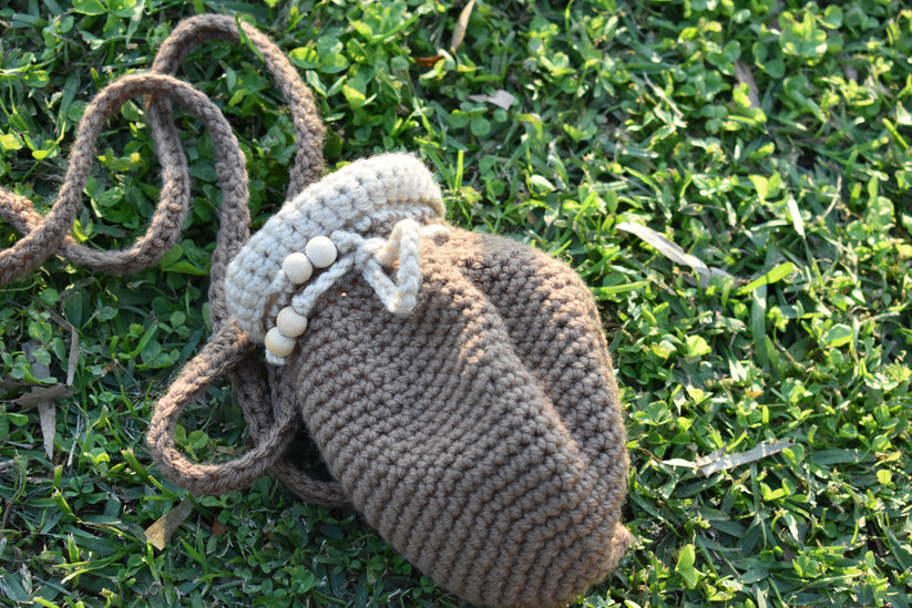 A handmade crocheted shoulder bag in taupe brown with a cream-colored beaded trim and strap, resting on a green grass background.