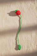 A handmade crochet toy designed like a strawberry, featuring a red body with green leafy stems and a long green tail, lying on a patch of grass outdoors.