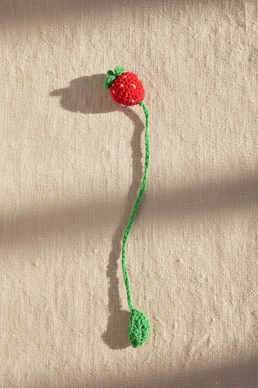 A handmade crochet toy designed like a strawberry, featuring a red body with green leafy stems and a long green tail, lying on a patch of grass outdoors.