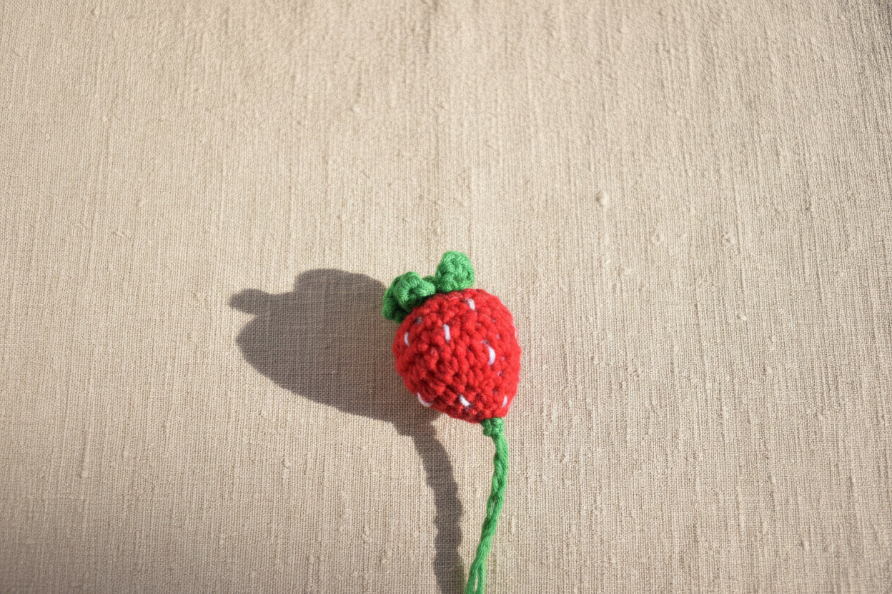 A small crocheted strawberry toy in red and green lies on grass outdoors.