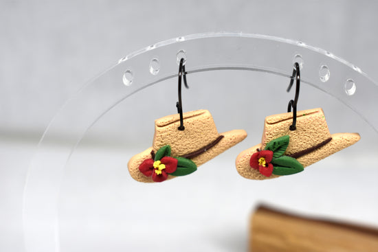 A pair of small cowboy hat earrings with red flowers and green leaves, hanging from black hooks on a clear acrylic display stand.