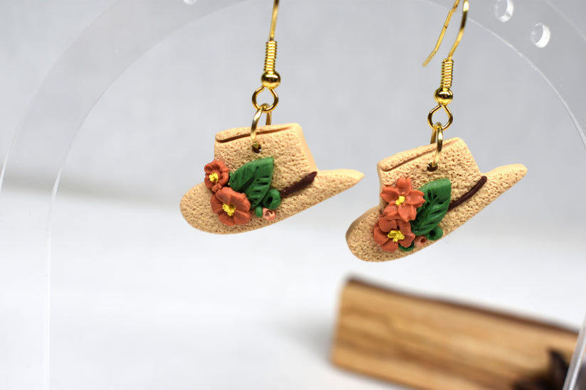 A pair of dangling earrings shaped like miniature straw cowboy hats, decorated with small orange flowers and green leaves, hanging from gold hooks against a soft white background.