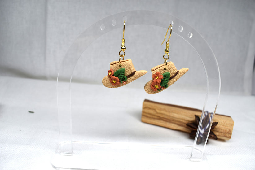 A pair of dangling earrings shaped like miniature straw cowboy hats, decorated with small orange flowers and green leaves, hanging from gold-tone hooks on a clear acrylic display stand.