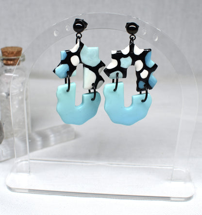 A pair of dangling earrings displayed on a clear acrylic stand. The earrings feature irregular light blue puffy shapes with black and white spotted upper sections and black posts.