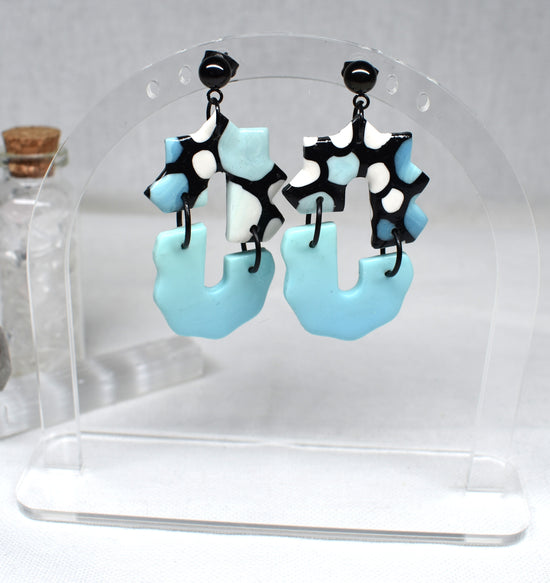 A pair of dangling earrings displayed on a clear acrylic stand. The earrings feature irregular light blue puffy shapes with black and white spotted upper sections and black posts.