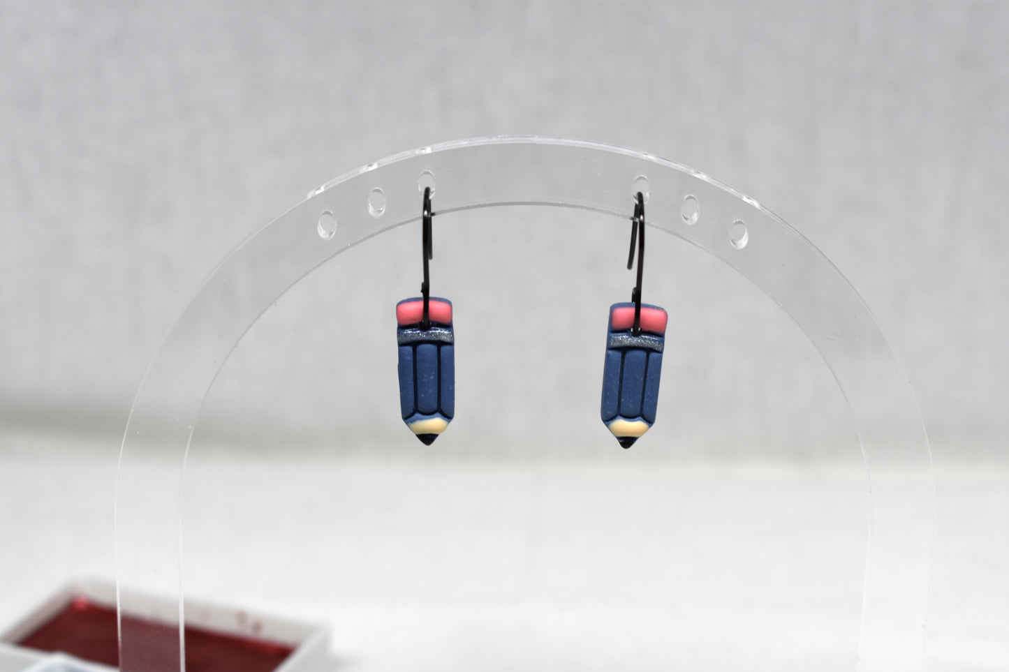 A pair of handmade polymer clay earrings shaped like pencils, featuring navy blue bodies, pink erasers, and gold-colored tips, displayed on a clear acrylic stand.