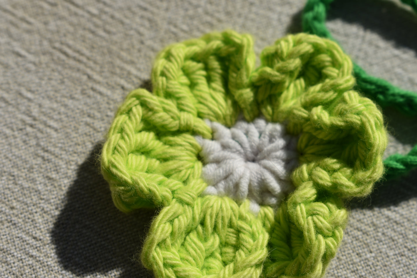 A close-up of a crocheted flower with lime green petals and a white center, resting on a textured beige fabric background.