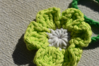A close-up of a crocheted flower with lime green petals and a white center, resting on a textured beige fabric background.